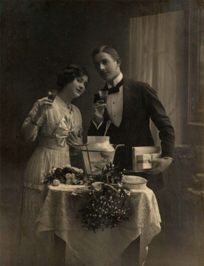 Пара з келихами в руках, біля накритого столу, оздобленого омелою, чоловік з подарунками. Фото 1900-1920 років.